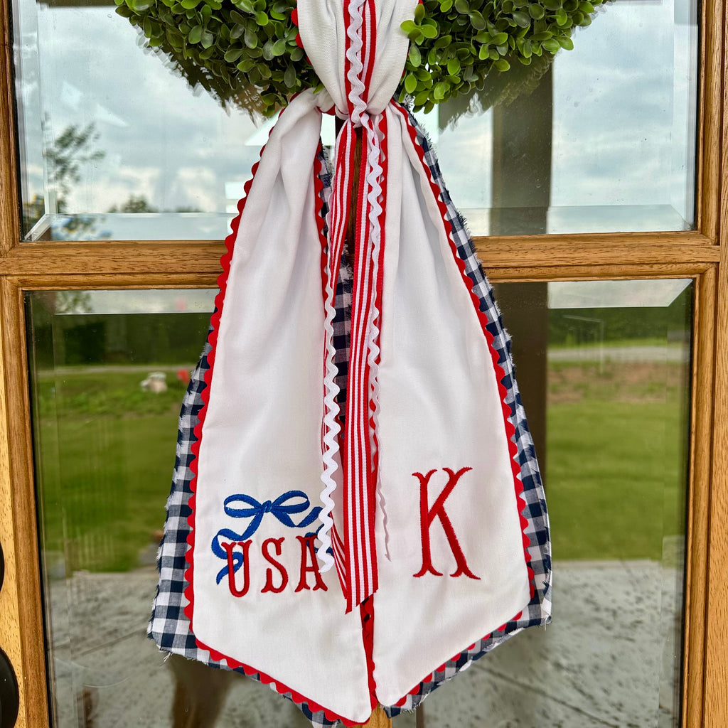 Decorative sash with 'USA' and 'K' monogram hanging on a door with a wreath in the background.
