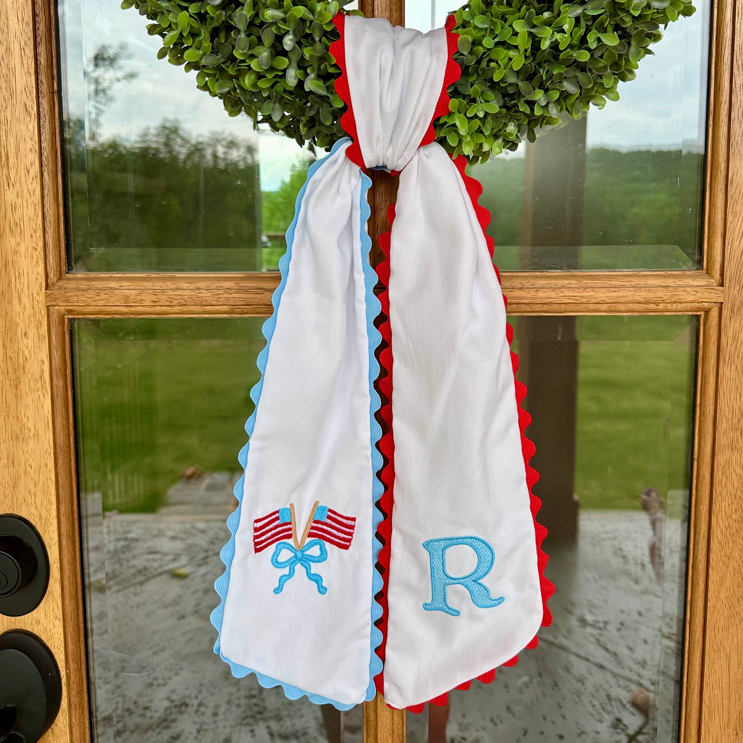 Decorative sash with embroidered letters and flags hanging on a door with green wreaths.