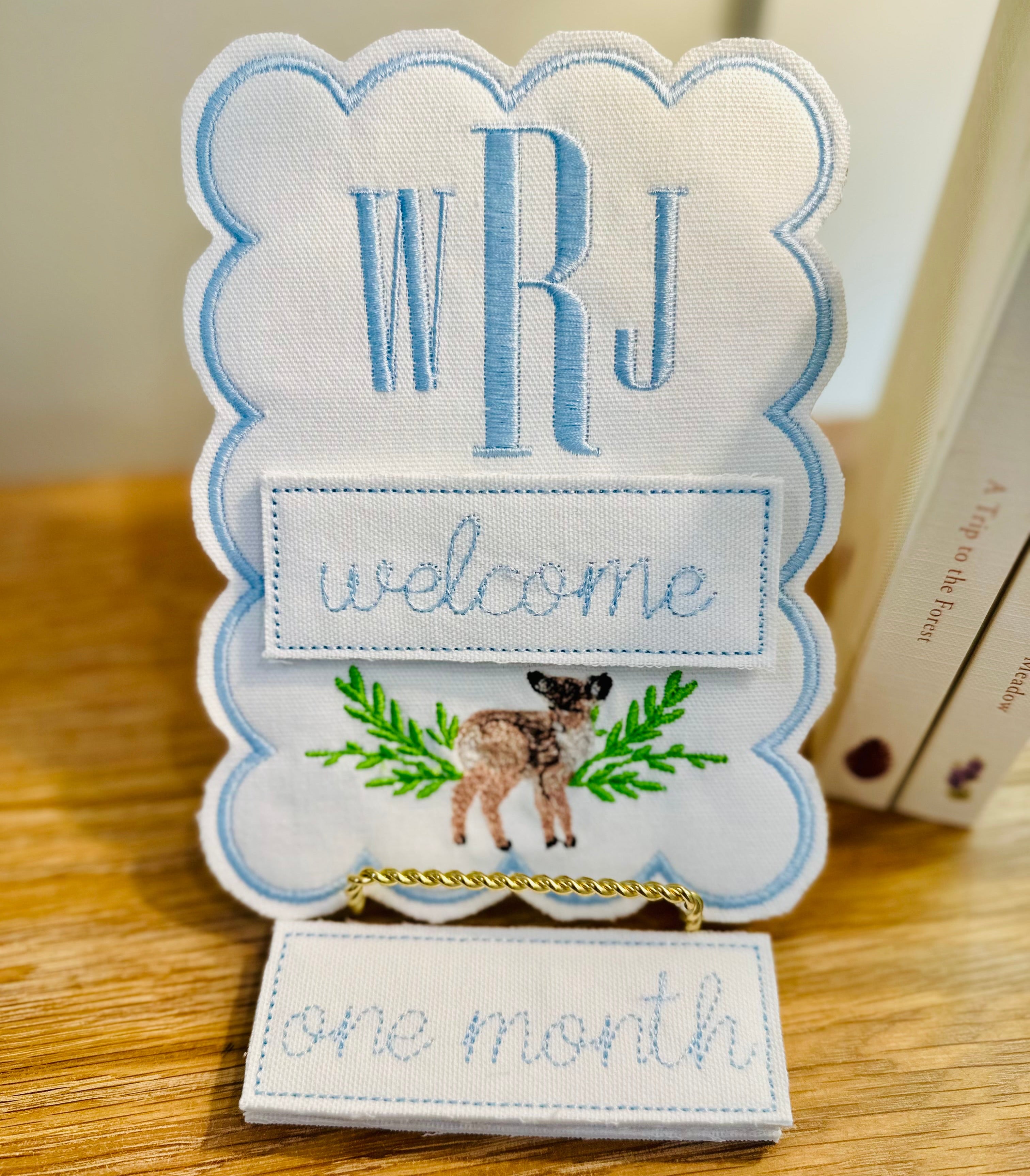 Embroidered card with 'WRJ welcome one month' text on a wooden surface
