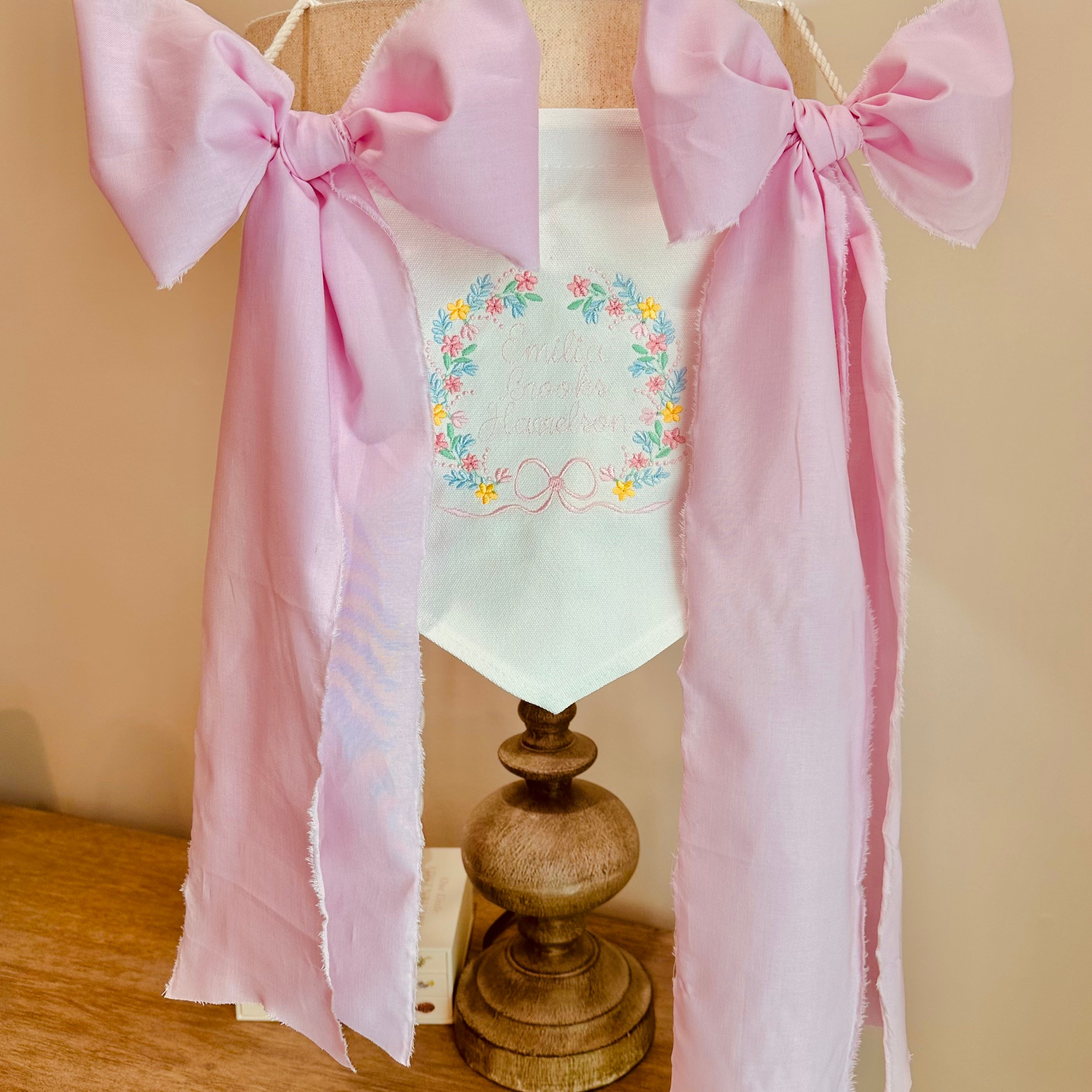 White baby banner with floral crest and lettering, has pink ribbon